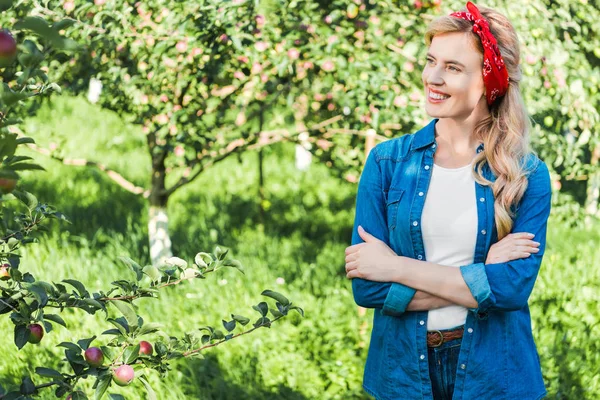 Sonriente agricultor atractivo de pie con los brazos cruzados en el jardín de manzanas - foto de stock