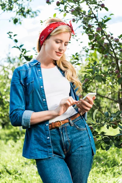 Привлекательный фермер с помощью смартфона в яблоневом саду на ферме — стоковое фото