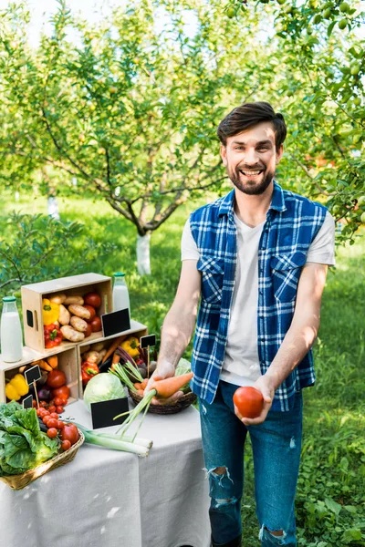 Apuesto granjero sonriente mostrando verduras ecológicas maduras en el mercado de granjeros - foto de stock