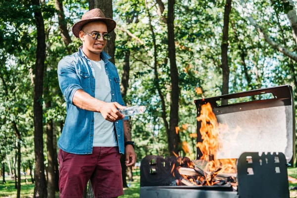 Smiling african american man in sunglasses and hat setting fire on grill in park — Stock Photo