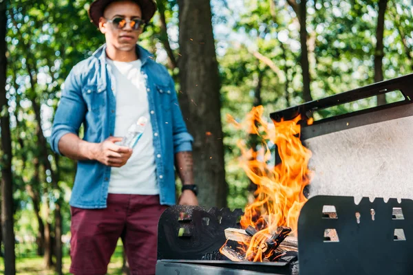Enfoque selectivo del hombre afroamericano en gafas de sol y sombrero prendiendo fuego a la parrilla en el parque - foto de stock
