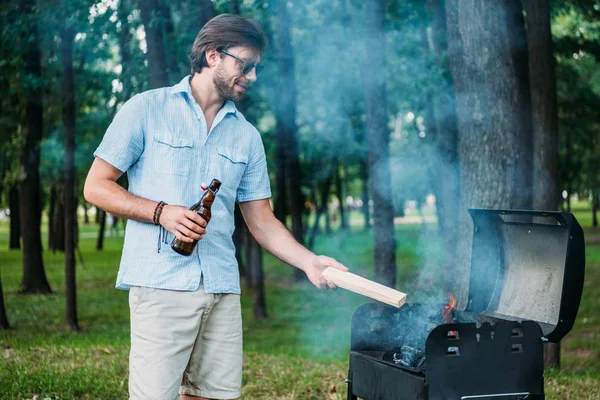 Smiling man in sunglasses with beer setting fire on grill during barbecue in park — Stock Photo