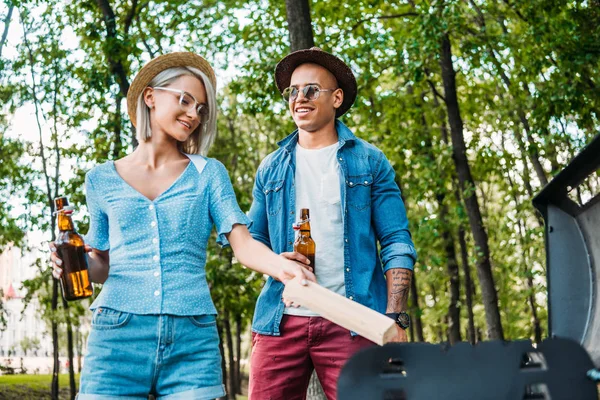 Multiracial cheerful couple with beer setting fire on grill in summer park — Stock Photo