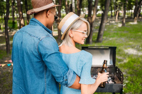 Vue arrière de l'homme afro-américain étreignant souriante petite amie caucasienne avec de la bière pendant le barbecue dans le parc — Photo de stock