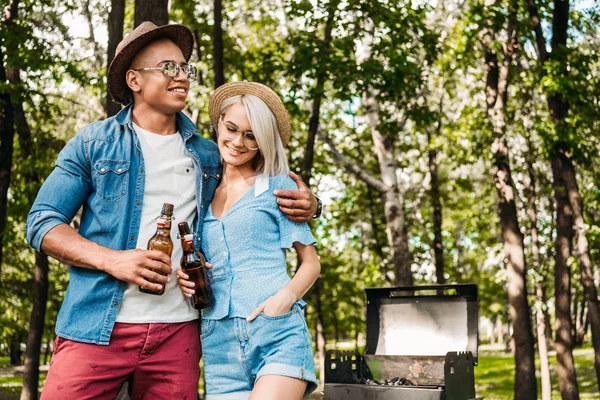 Retrato de pareja multirracial con cerveza de pie cerca de la parrilla en el parque de verano - foto de stock