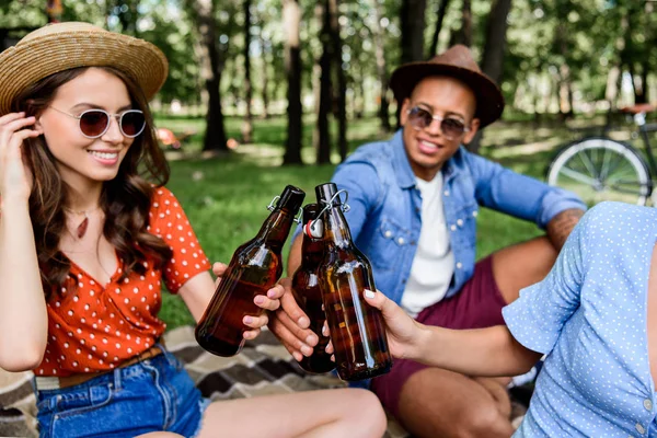 Partial view of interracial friends clinking bottles of beer during picnic in park — Stock Photo