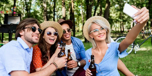 Souriant amis multiculturels avec la bière prendre selfie sur smartphone tout en se reposant dans le parc d'été — Photo de stock