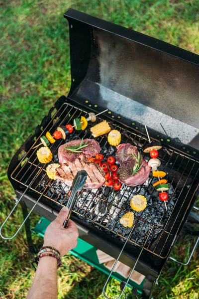 Cropped shot of man with tongs cooking food on grill during barbecue in park — Stock Photo