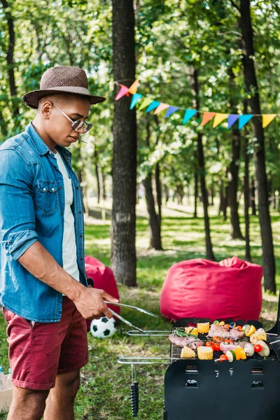Side view of african american man in sunglasses and hat cooking food on grill in park — Stock Photo
