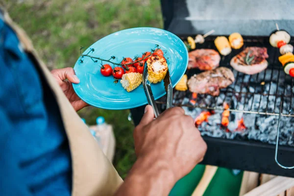 Teilansicht eines afrikanisch-amerikanischen Mannes mit einer Zange, die gegrilltes Gemüse während des Grillens im Park auf den Teller legt — Stockfoto