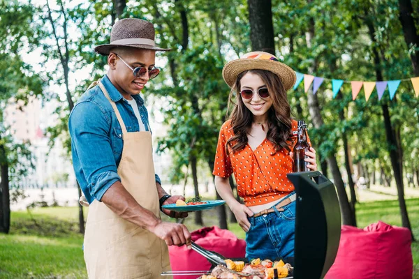 Donna sorridente con birra guardando il fidanzato afro-americano che cucina cibo sulla griglia durante il barbecue — Foto stock