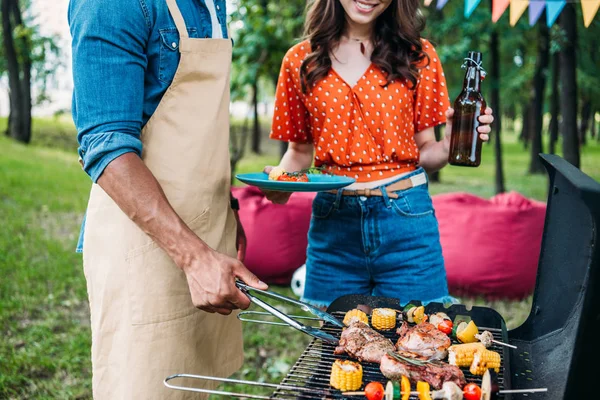 Vista parziale della donna con birra guardando il fidanzato afro-americano che cucina cibo alla griglia durante il barbecue — Foto stock