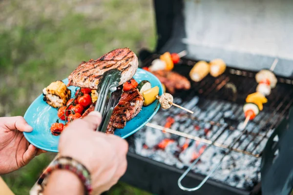 Visão parcial do homem com pinças colocando comida grelhada na placa durante bbq no parque — Fotografia de Stock