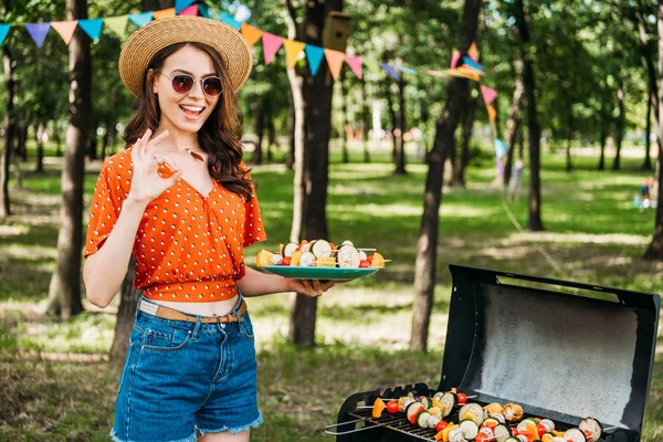 Портрет счастливой женщины в шляпе и солнцезащитных очках с овощами на тарелке, показывающий знак ОК в парке — стоковое фото
