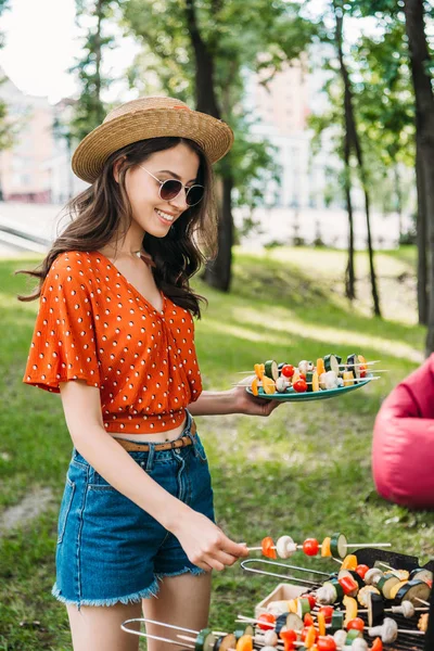 Seitenansicht einer lächelnden jungen Frau mit Hut und Sonnenbrille, die im Park Gemüse vom Grill holt — Stockfoto