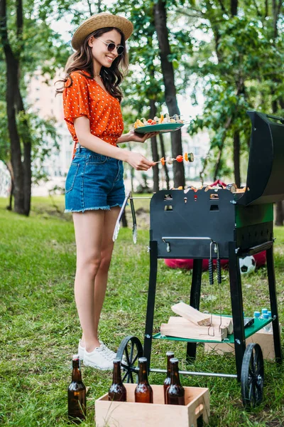 Вид сбоку улыбающейся молодой женщины, которая берет овощи из гриля в парке — стоковое фото