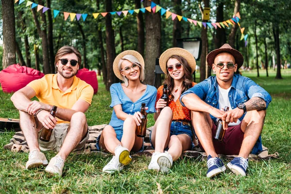 Счастливые многорасовые друзья с пивом, отдыхающие на одеяле в летнем парке — стоковое фото