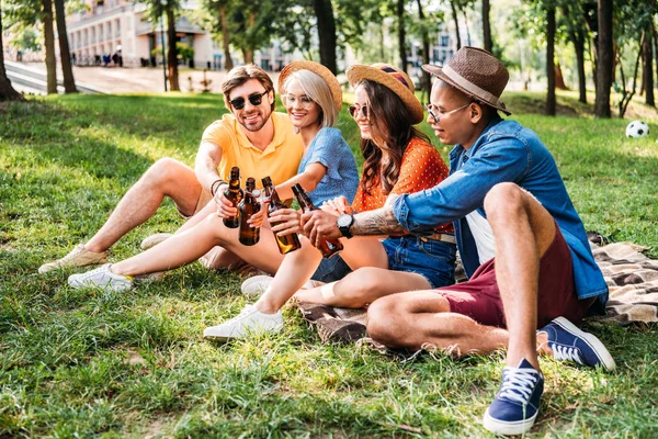 Щасливі багаторасові друзі смердять пляшки пива, відпочиваючи на ковдрі в літньому парку — стокове фото