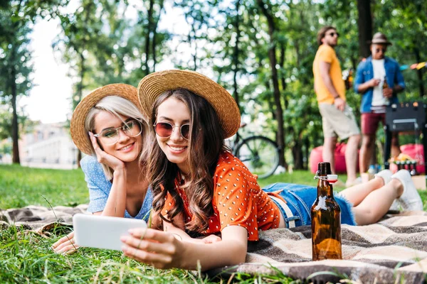Selective focus of smiling friends taking selfie on smartphone while resting on blanket during bbq in park — Stock Photo