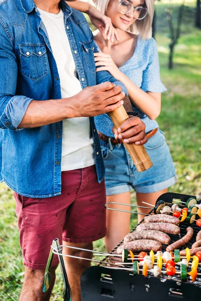 Vista parcial de la mujer de pie cerca de novio afroamericano mientras que él cocina comida en la parrilla - foto de stock