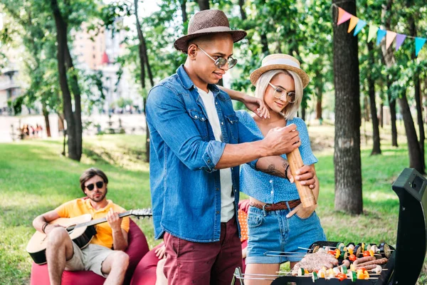 Mujer de pie cerca de afroamericano novio mientras él cocinar comida en la parrilla - foto de stock