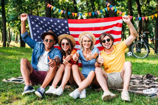 Amigos multirraciales felices con bandera americana mostrando pulgares hacia arriba en el parque de verano - foto de stock