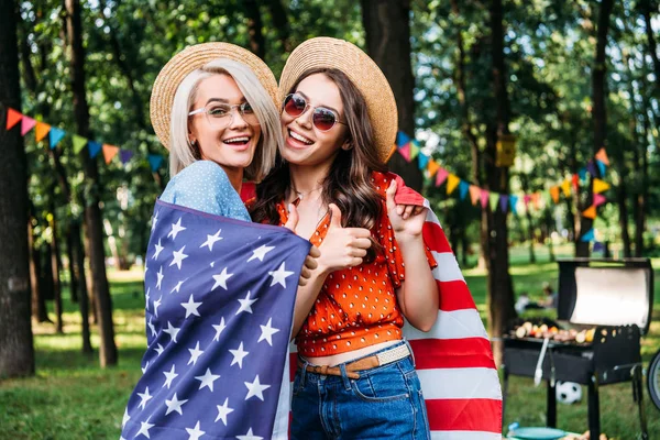 Retrato de mujeres felices en sombreros y gafas de sol con bandera americana en el parque - foto de stock