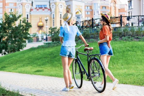 Vista trasera de las mujeres jóvenes en sombreros con bicicleta caminando por la calle - foto de stock