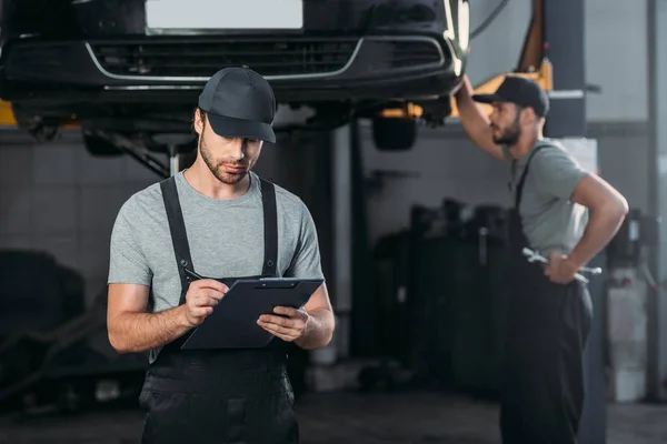 Mecánico de automóviles en overoles escribir en portapapeles, mientras que el colega que trabaja en el taller detrás — Stock Photo