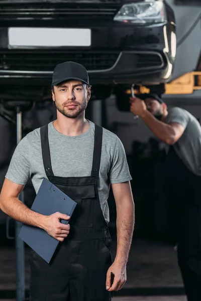 Mecánico profesional en overoles sujetando portapapeles, mientras que su colega trabaja en el taller detrás - foto de stock