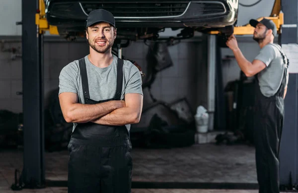 Lächelnder Mechaniker in Overalls posiert mit verschränkten Armen, während sein Kollege in der Werkstatt hinter ihm arbeitet — Stockfoto