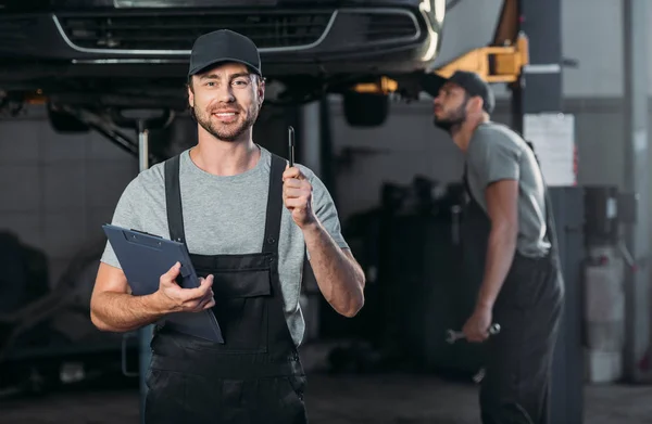 Smiling mechanic wirh clipboard having idea, while colleague working in workshop behind — Stock Photo