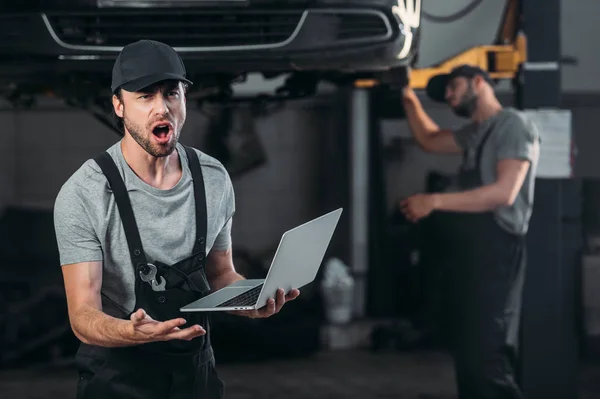Schockierter Mechaniker mit Laptop, während Kollege in Werkstatt dahinter arbeitet — Stockfoto