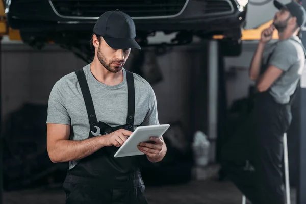 Mecânico automóvel profissional em macacão usando tablet digital, enquanto colega de trabalho na oficina atrás — Fotografia de Stock