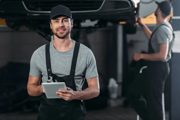 Mecánico feliz en overoles usando tableta digital, mientras que colega que trabaja en taller detrás - foto de stock