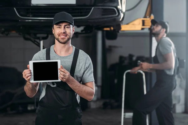 Automechaniker hält digitales Tablet mit leerem Bildschirm in der Hand, während Kollege im Hintergrund in der Werkstatt arbeitet — Stockfoto