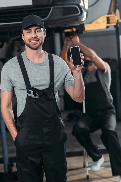Mecánico automático que muestra el teléfono inteligente con pantalla en blanco, mientras que el colega que trabaja en el taller detrás - foto de stock