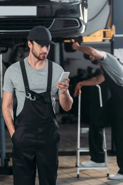 Professional auto mechanic using smartphone, while colleague working in workshop behind — Stock Photo
