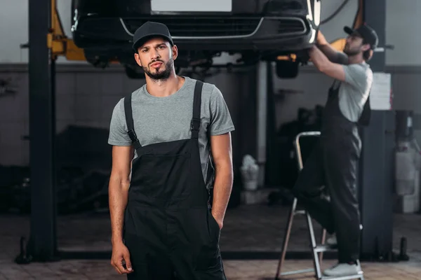 Automechaniker posiert in Overalls, während Kollege in Werkstatt dahinter arbeitet — Stockfoto