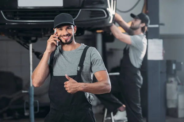 Mecánico automático hablando en el teléfono inteligente y mostrando el pulgar hacia arriba, mientras que el colega que trabaja en el taller detrás - foto de stock