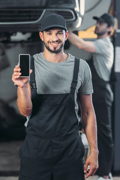 Mecánico profesional que muestra el teléfono inteligente con pantalla en blanco, mientras que el colega que trabaja en el taller detrás - foto de stock