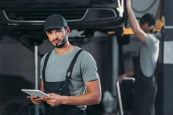 Mecánico de automóviles en overoles utilizando tableta digital, mientras que el colega que trabaja en el taller detrás — Stock Photo