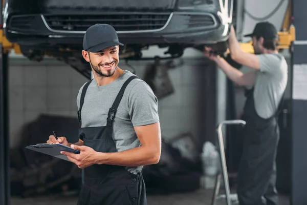 Professional mechanic writing in clipboard, while colleague working in workshop behind — Stock Photo