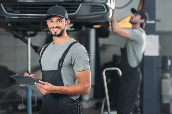 Escritura mecánica sonriente en portapapeles, mientras que colega que trabaja en el taller detrás - foto de stock