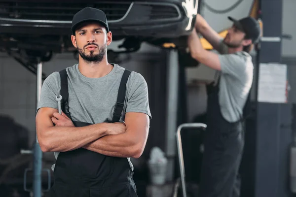 Seriöser Mechaniker mit verschränkten Armen, der Schraubenschlüssel hält, während der Kollege in der Werkstatt hinter ihm arbeitet — Stockfoto