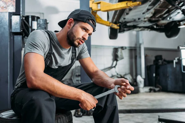 Mecánico cansado sosteniendo la llave inglesa y sentado en taller de reparación de automóviles — Stock Photo