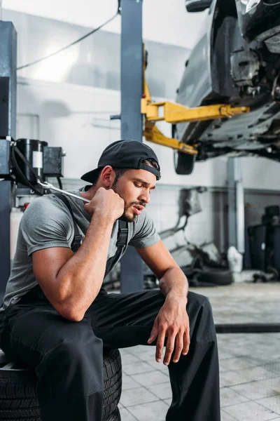 Tired auto mechanic holding wrench and sitting in repair shop — Stock Photo