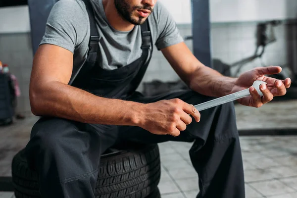 Cropped view of professional mechanic holding wrench and sitting in auto repair shop — Stock Photo