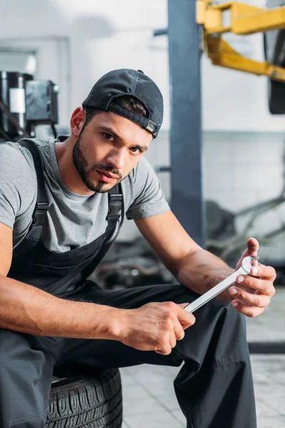 Profi-Mechaniker hält Schraubenschlüssel und sitzt in Autowerkstatt — Stockfoto