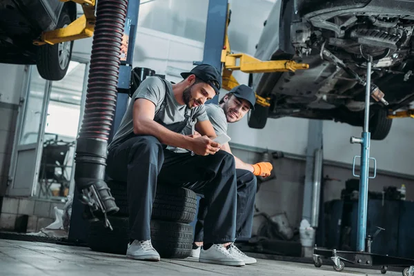Two auto mechanics using smartphone and resting in auto repair shop — Stock Photo